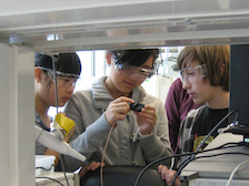 three young students wearing goggles examine a device at a lab bench