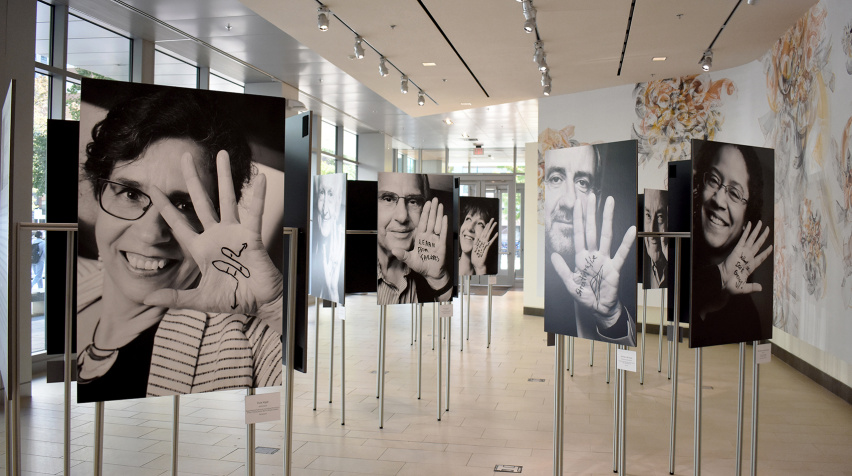 Black and white portraits mounted on metal posts fill an open space with lights around them. Each scientist raises a hand, on which a phrase or science illustration is drawn
