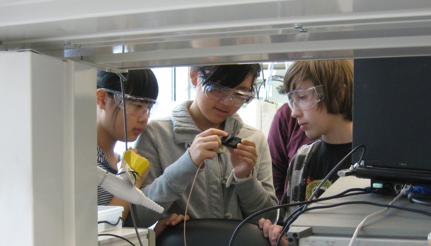 Three young people interact with a small device at a lab bench