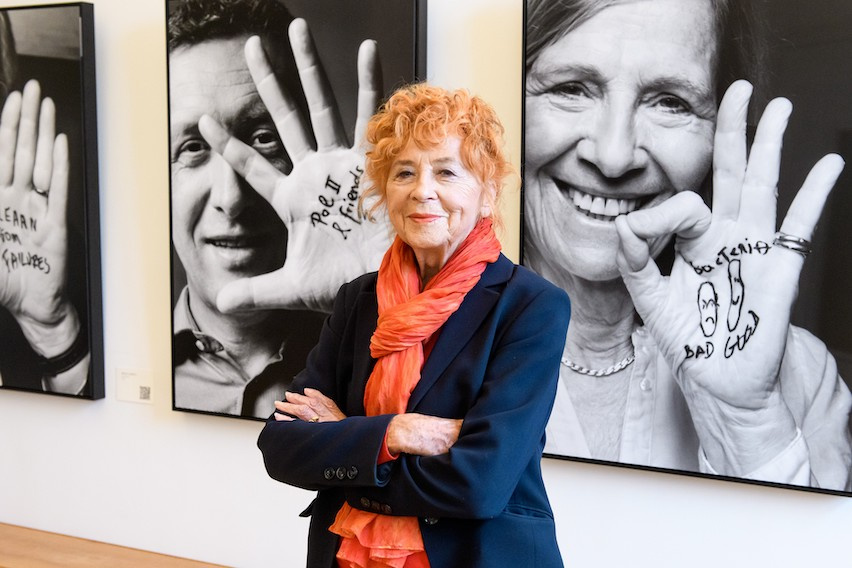 Redheaded photographer Herlinde Koelbl stands with her arms crossed, photographic portraits of scientists behind her