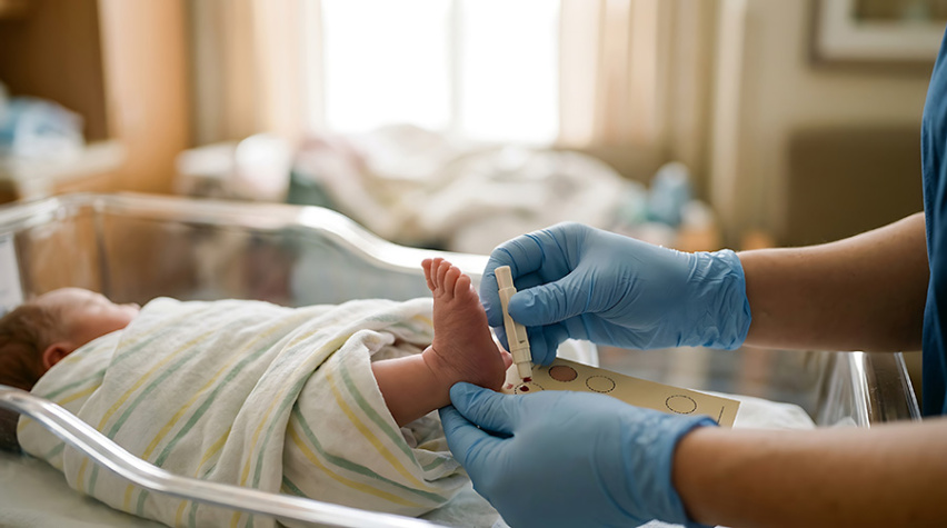 A nurse holds a newborn's foot, dropping a small amount of blood from a heel stick onto a paper