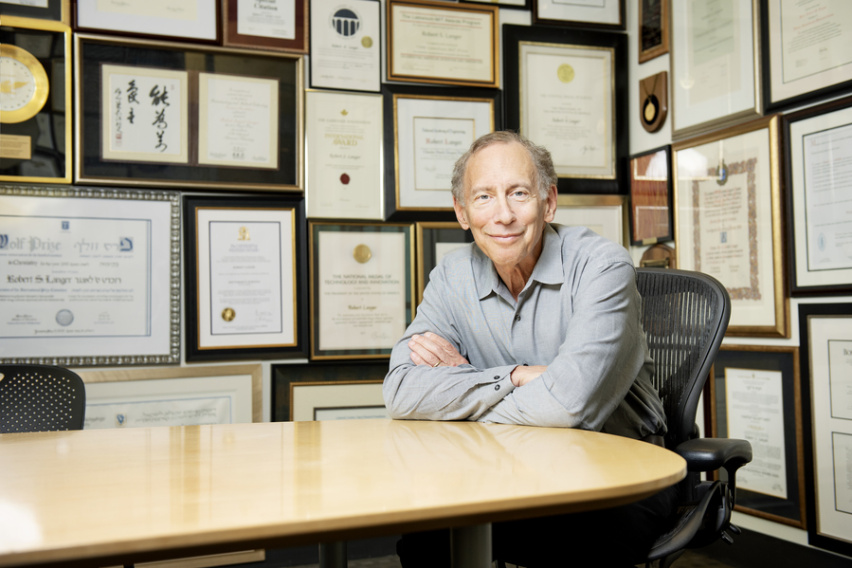 Robert Langer seated at a table with a wall of framed awards behind