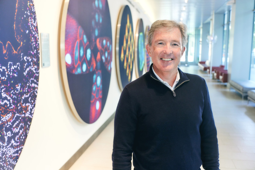 Photo of Tyler Jacks standing in front of a wall of colorful scientific images in the Koch Institute lobby and smiling for the camera.