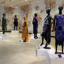ten black matte mannequins draped in colorful fabrics stand on pedestals in a brightly lit atrium