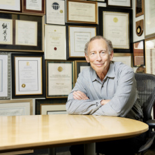 Robert Langer seated at a table with a wall of framed awards behind