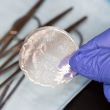 A gloved hands holds a circular semi-transparent film above a tray of scissors