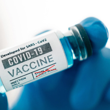 A close-up shot of a vial with the words "COVID-19 vaccine" being held by a hand covered in a blue glove