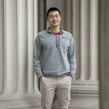 Photo of Alex Tang, wearing a grey sweatshirt and standing in front of a building on the MIT campus.