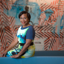 Photo of Paula Hammond, wearing a blue, green, yellow and white dress and smiling for the camera.  