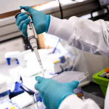 Close-up of the arms of scientist in white lab coat wearing blue gloves and holding an injection device.