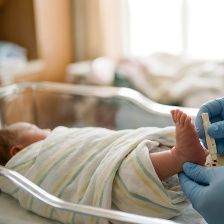 Photo of a baby swathed in a blanket with it's foot sticking out while a pair of blue-gloved hands administers a heel stick test
