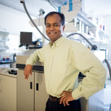 Photo of Ram Sasisekharan wearing a pale yellow shirt standing in his laboratory and smiliing for the camera 