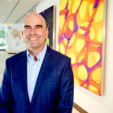 Photo of Forest White, wearing a navy blue suit, standing in front of a wall of colorful scientific images in the Koch Institute lobby.