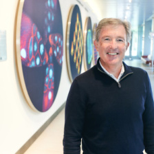 Photo of Tyler Jacks standing in front of a wall of colorful scientific images in the Koch Institute lobby and smiling for the camera. 