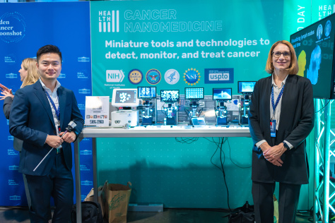 Ana Jaklenec and Jeremy Li stand in front of their exhibition booth, which includes several devices with view screens.