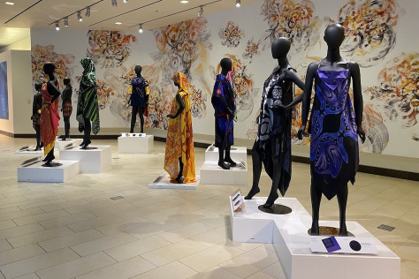ten black matte mannequins draped in colorful fabrics stand on pedestals in a brightly lit atrium