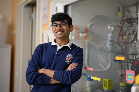 A young man in a rugby shirt stands arms crossed in a lab