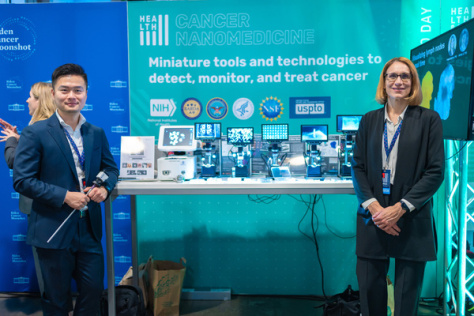 Ana Jaklenec and Jeremy Li stand in front of their exhibition booth, which includes several view screens.