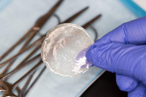 A gloved hand holds a quasi-transparent circular film over a tray of metal instruments