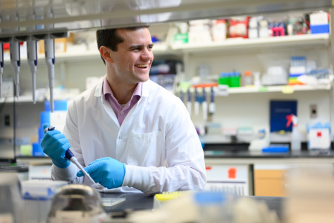 Louis DeRidder in a lab, wearing a white coat and gloves and holding a piece of equipment.