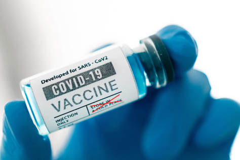 A close-up shot of a vial with the words "COVID-19 vaccine" being held by a hand covered in a blue glove