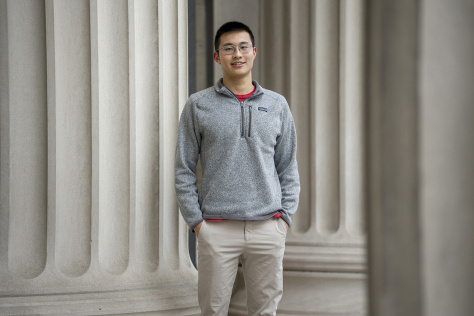 Photo of Alex Tang, wearing a grey sweatshirt and standing in front of a building on the MIT campus. 