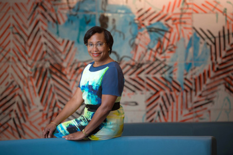 Photo of Paula Hammond, wearing a blue, green, yellow and white dress and smiling for the camera.  