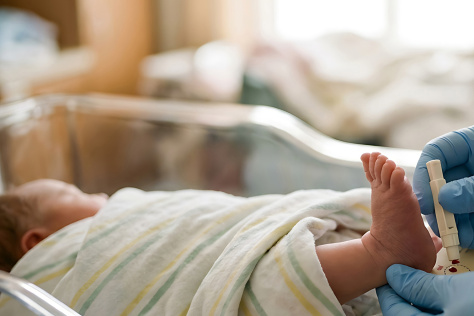 Photo of a baby swathed in a blanket with it's foot sticking out while a pair of blue-gloved hands administers a heel stick test