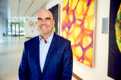 Photo of Forest White, wearing a navy blue suit, standing in front of a wall of colorful scientific images in the Koch Institute lobby.