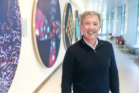 Photo of Tyler Jacks standing in front of a wall of colorful scientific images in the Koch Institute lobby and smiling for the camera. 