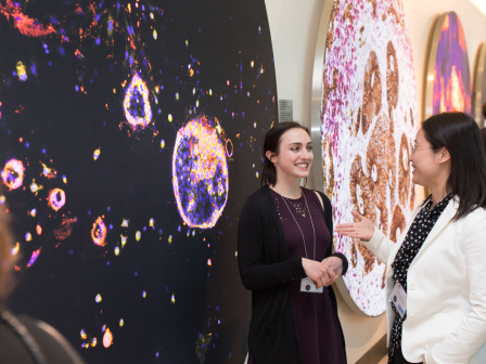 Two women standing in front of a wall of scientific images, talking and smiling. 