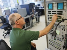 A seated man presses a button on a tall machine with two screens and many buttons and toggles