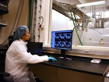 A woman in lab safety gear sits at a computer in front a window overlooking large machines.